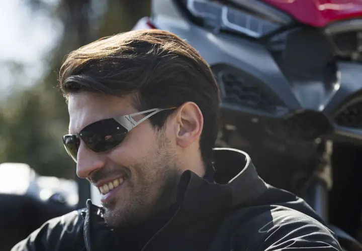 Uomo sorridente con occhiali da moto sportivi, con una moto sullo sfondo.