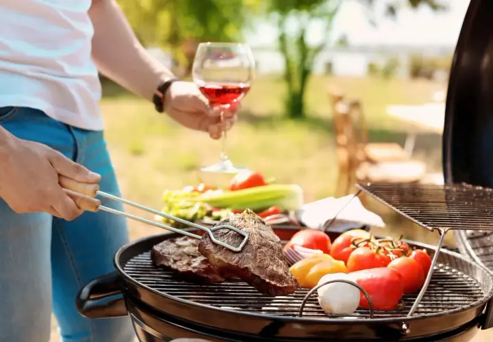 Un homme savoure un verre de vin rouge tout en préparant un barbecue avec des steaks et des légumes.