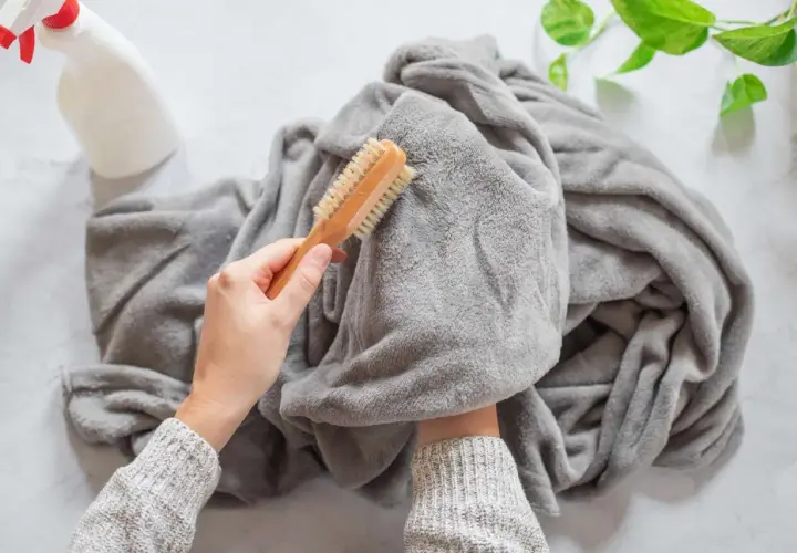 A person is brushing a grey throw blanket, showing how to wash it. A spray bottle is nearby.