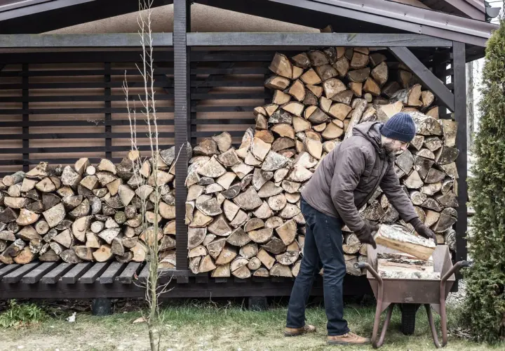 Mann mit blauer Mütze lagert Holz in einer Schubkarre vor einem Holzschuppen.