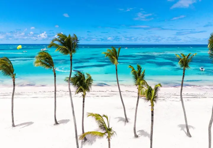 Traumhafter **Urlaub im November** an einem karibischen Strand mit Palmen, weißem Sand und türkisfarbenem Meer. Boote und Parasailing im Hintergrund.