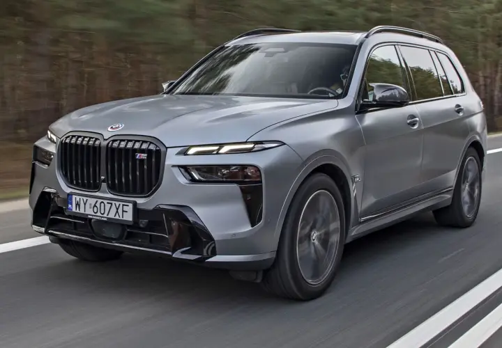 Szary BMW X7 2025 z czarnym grillem i sportowymi akcentami pędzi po drodze wśród drzew.