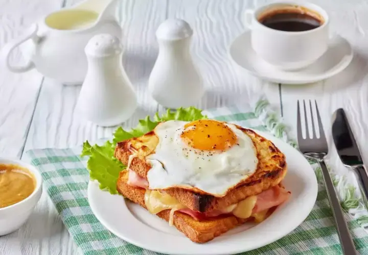 Un délicieux croque monsieur garni d'un œuf au plat, servi avec une tasse de café et une sauce crémeuse.