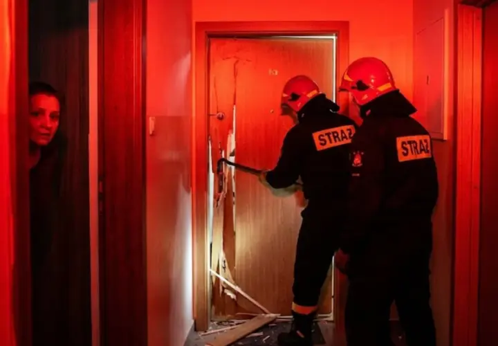 Strażacy wyważają drzwi, gdy kobieta patrzy z niepokojem. Sytuacja, kiedy policja może wyważyć drzwi, by ratować życie lub zapobiec przestępstwu.