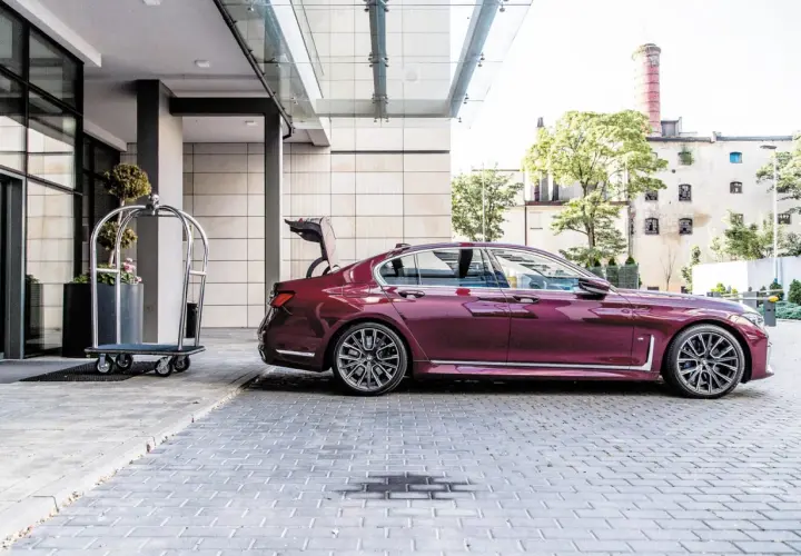 Eleganckie bmw g11 w kolorze śliwkowym z otwartym bagażnikiem stoi przed hotelem obok wózka na bagaż.