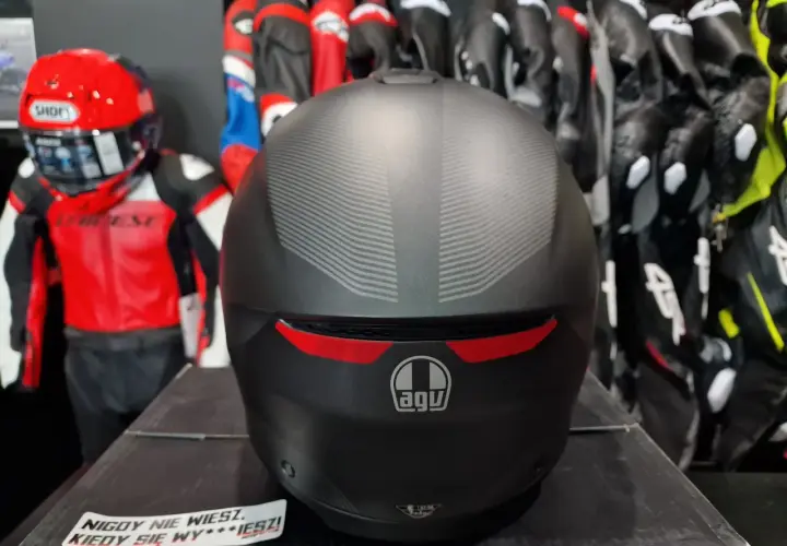 Matowy kask AGV z subtelnym wzorem, gotowy na przygodę. W tle czerwono-czarny kombinezon motocyklowy.