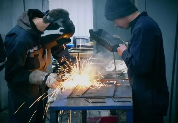 Dwóch spawaczy pracuje przy metalowej konstrukcji. Jeden spawa elektrodą, tworząc snop iskier. Drugi obserwuje, trzymając narzędzie.