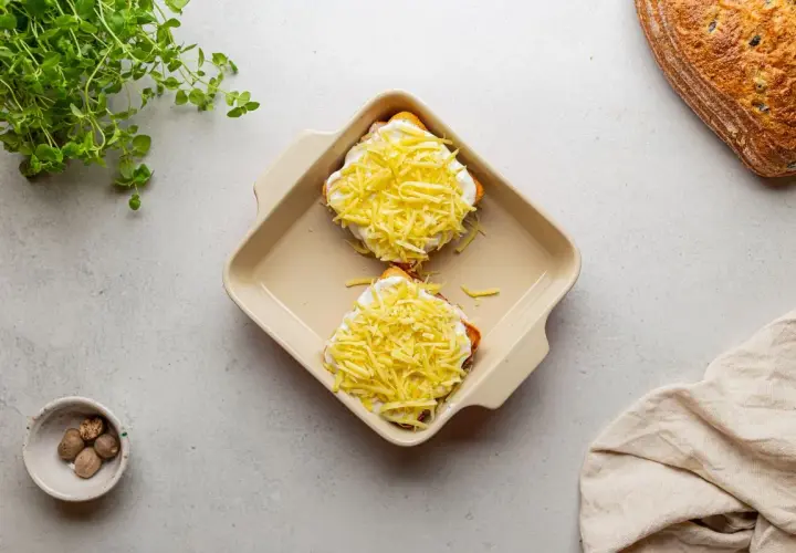 Dwa tosty z szynką i serem, przygotowane do zapieczenia, jak w klasycznym croque madame. Obok gałka muszkatołowa i świeża zielenina.