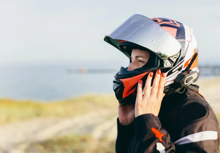 Donna con casco da moto colorato, con mani che aggiustano la visiera per evitare che si appanni. Sfondo sfocato di mare e sabbia.