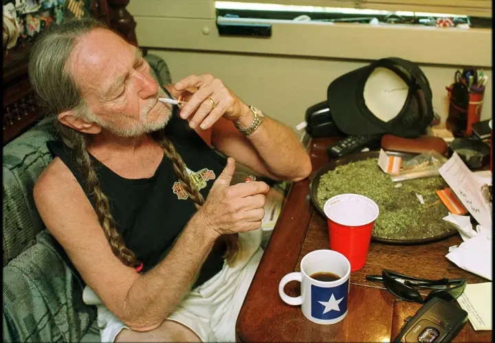 Willie Nelson, icona delle canzoni country famose, si accende una sigaretta con una tazza di caffè accanto.
