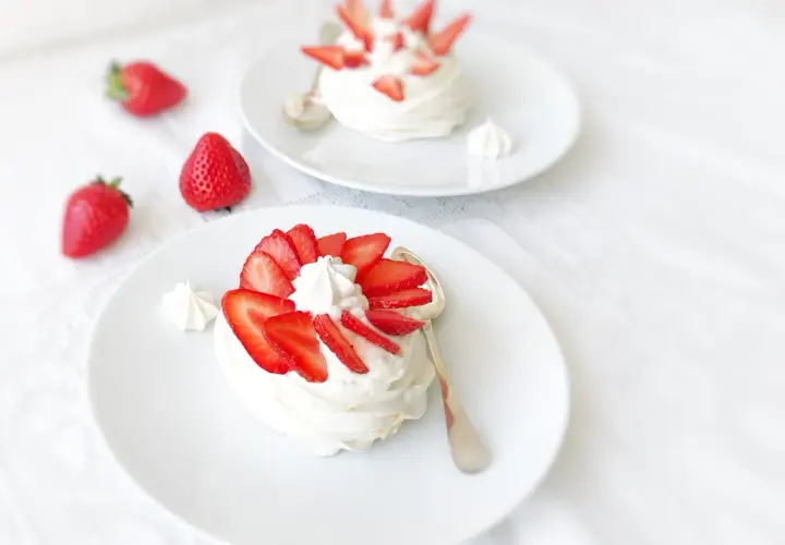 Deux pavlovas à la fraise, légers et aériens, ornés de fruits rouges frais et de crème fouettée, sur des assiettes blanches.