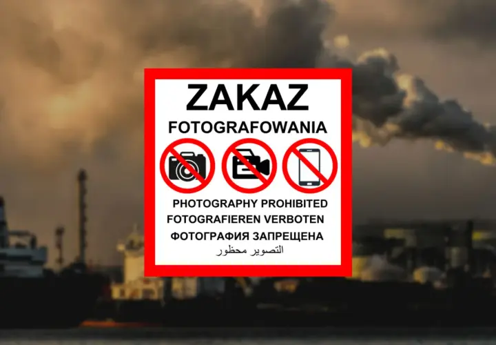 Tablica z napisem "ZAKAZ FOTOGRAFOWANIA" i symbolami przekreślonych aparatów, kamer i telefonów komórkowych. W tle widać przemysłowy krajobraz z kominami fabrycznymi i statkami. To znak informujący, czego nie można fotografować w Polsce.