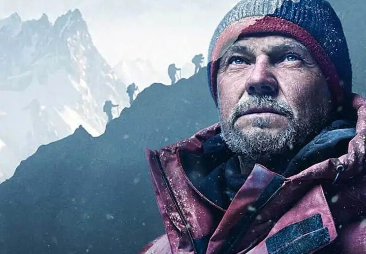 Zmęczony alpinista w czapce i kurtce patrzy w górę. W tle widać innych wspinaczy na zboczu podczas wyprawy na Broad Peak.