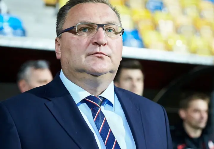 Selekcjoner w okularach, w garniturze i krawacie, z uwagą obserwuje wydarzenia na boisku.