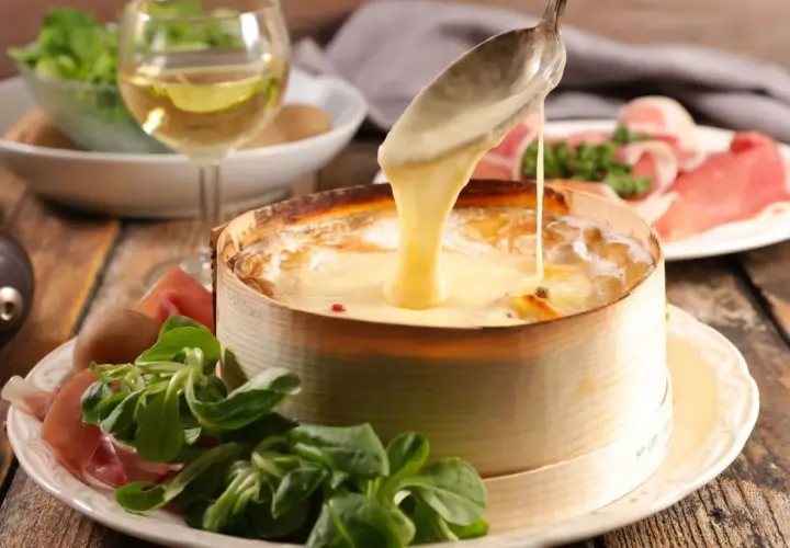 Un Mont d'Or fondant est servi avec une cuillère, accompagné de charcuterie, de pommes de terre et d'une salade verte. Un verre de vin blanc complète ce plat gourmand.