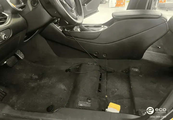 Interior de coche con moqueta oscura y algo de suciedad. Se ve el volante, consola central y pedales. Hay cables y un pequeño objeto amarillo.