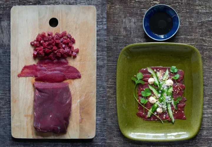 Förberedelser för asiatisk råbiff: rått kött i bitar och skivor på en skärbräda, färdig rätt med garnering på en grön tallrik.