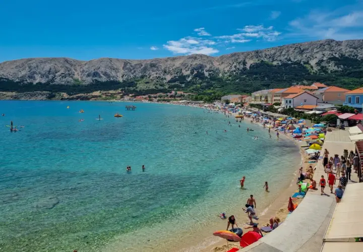 Słoneczna plaża na wyspie Krk, pełna ludzi kąpiących się i opalających. W tle góry i zabudowania. To idealne miejsce na wakacyjne atrakcje.