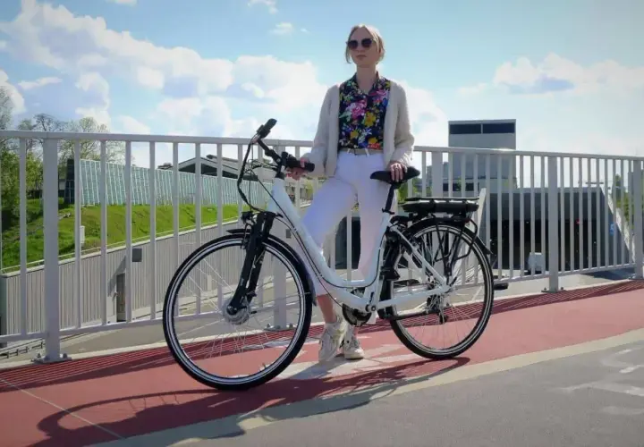 Kobieta w białych spodniach i kolorowej koszuli stoi obok białego roweru elektrycznego. Czy na rower elektryczny trzeba mieć prawo jazdy?