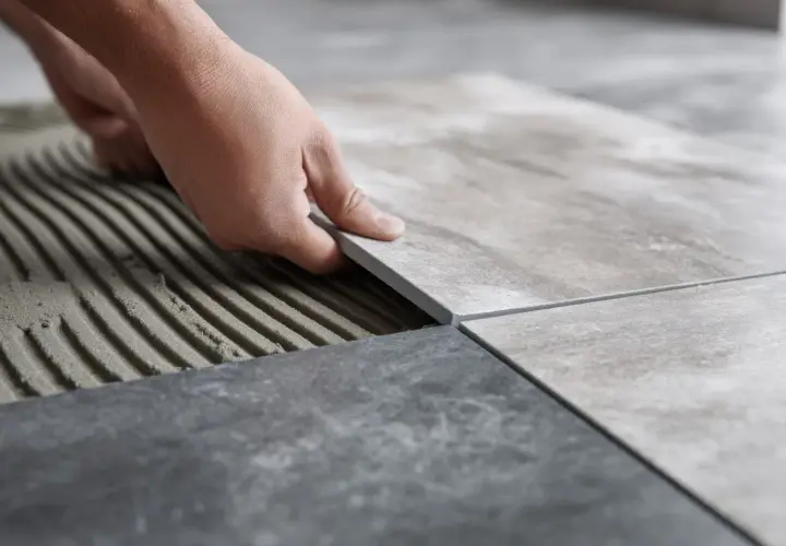 Układanie płytek 60x60. Cennik prac glazurniczych. Dłoń kładzie nową płytkę na kleju.