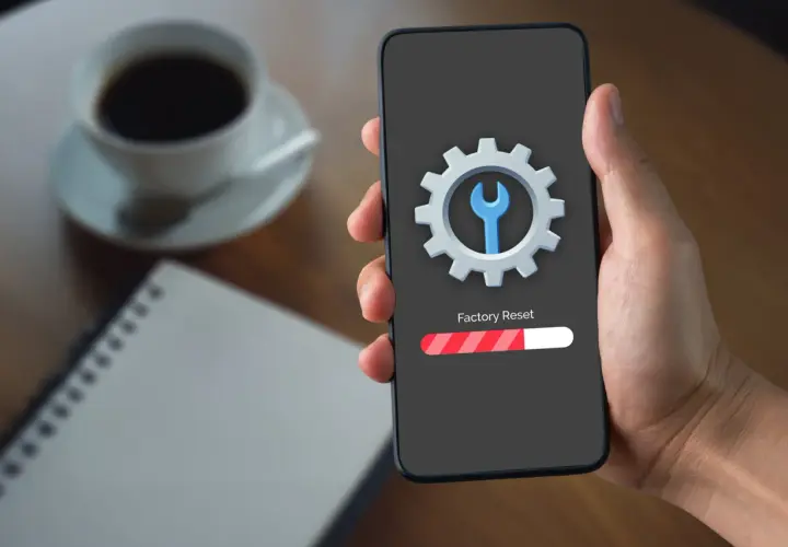 Dłoń trzyma smartfon z ekranem pokazującym ikonę koła zębatego i klucza, oraz pasek postępu "Factory Reset". To rozwiązanie, jak zresetować telefon gdy nie działa dotyk.