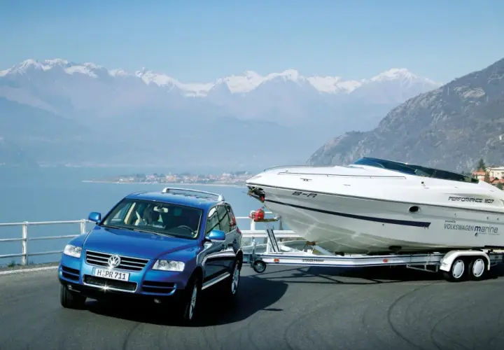 Un coche azul remolca un barco deportivo blanco por una carretera junto a un lago y montañas nevadas.