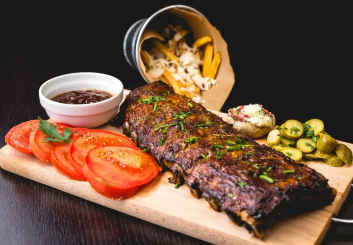 Un plat de côtes de porc au barbecue, garni de ciboulette, servi avec des tomates tranchées, des cornichons, une sauce barbecue et des frites au fromage.