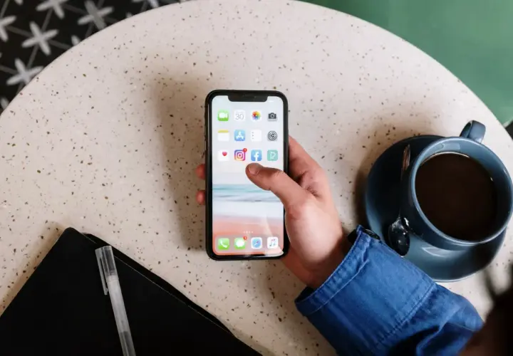 Main d'une personne tenant un smartphone affichant des icônes d'applications, comme un exemple d'application web. Une tasse de café est à côté.