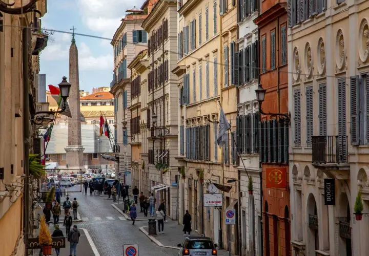 Una strada romana con palazzi storici, un obelisco e gente che passeggia. Si intuisce quanto è lunga Via del Corso, con la sua prospettiva che si perde all'orizzonte.