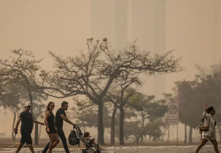 Gente camina bajo un cielo anaranjado, reflejo de las causas de la contaminación del aire. Árboles secos y edificios difuminados completan la escena.