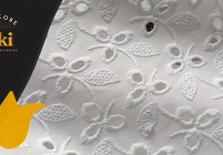 Close-up of white eyelet fabric with floral embroidery. This delicate fabric features cut-out holes surrounded by decorative stitching, creating a beautiful, airy pattern.
