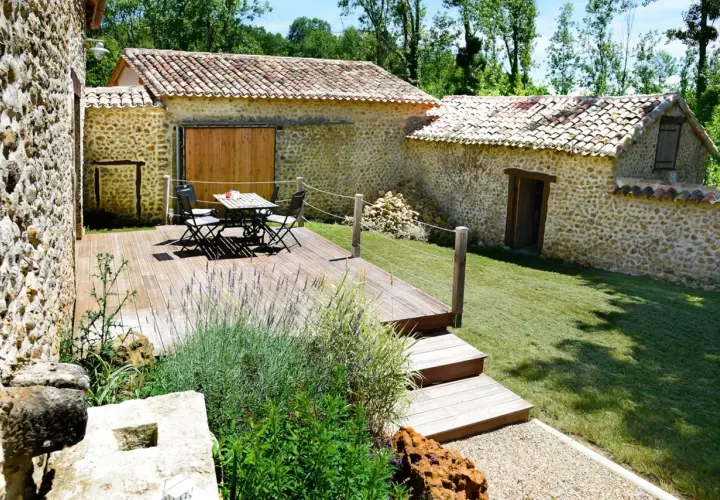 Aménagement longère : terrasse en bois avec mobilier, jardin verdoyant et murs en pierre.