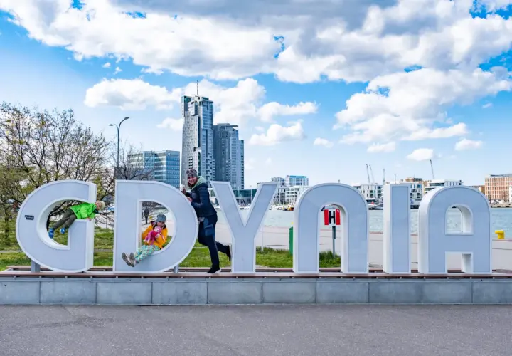 Dzieci bawią się na literach napisu "GDYNIA" na tle miejskiego krajobrazu.