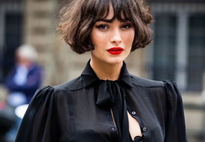 Mujer con un elegante corte de pelo **french bob**, labios rojos vibrantes y una blusa negra transparente con lazada.