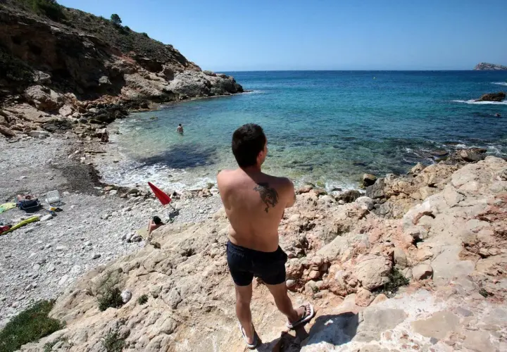 Hombre con tatuaje en la espalda disfruta de la vista en la playa nudista Benidorm. Aguas cristalinas y rocas caracterizan este rincón.