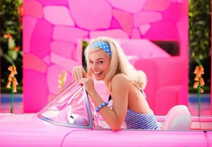 Margot Robbie jako Barbie w różowym kabriolecie, z uśmiechem pozuje na tle jaskrawego, różowego świata.