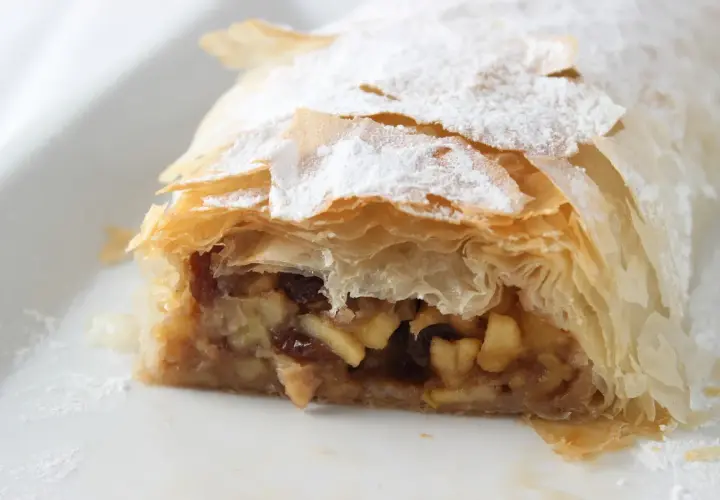 Un trozo de strudel de manzana, con su masa hojaldrada y relleno de manzana y pasas, espolvoreado con azúcar glas.