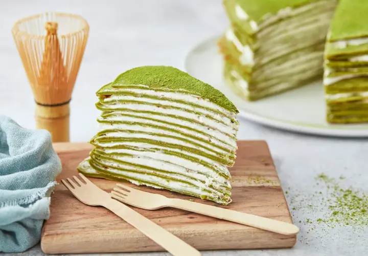 Ein Stück saftiger Matcha Kuchen mit vielen dünnen Schichten und cremiger Füllung, bestäubt mit grünem Pulver. Daneben ein Chasen und Holzgabeln.