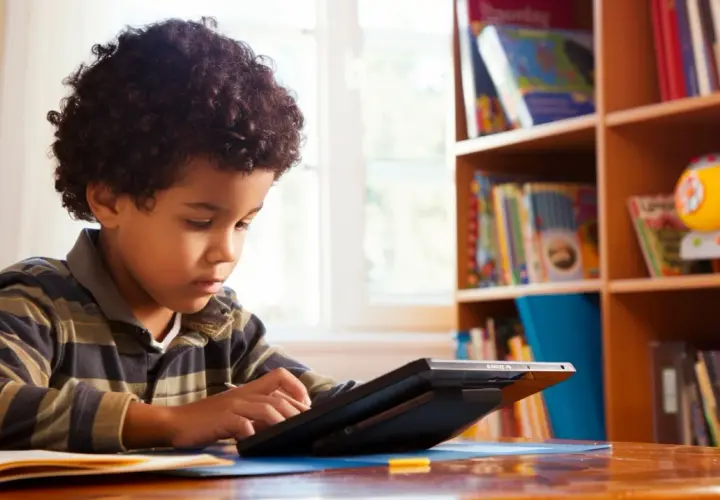 Ein Junge mit lockigem Haar lernt mit Bildungs-Apps auf einem Tablet. Im Hintergrund ein Bücherregal voller Bücher.