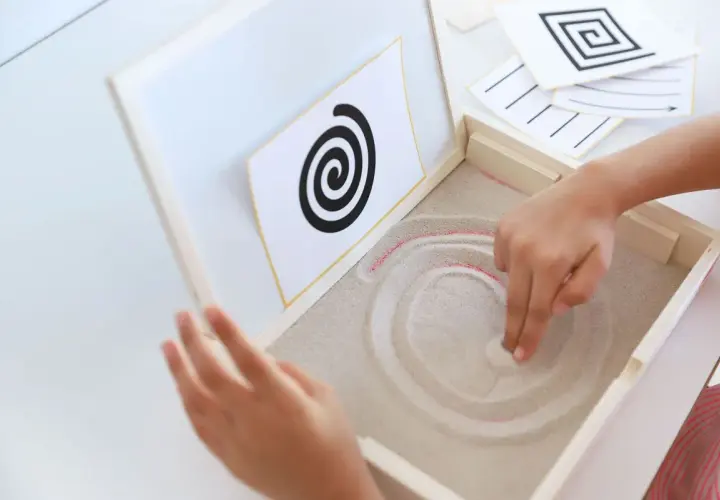Niño practicando preescritura montessori, trazando espirales en arena.