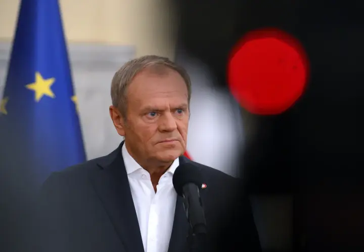 Donald Tusk w garniturze, z flagą UE w tle i czerwonym światłem.
