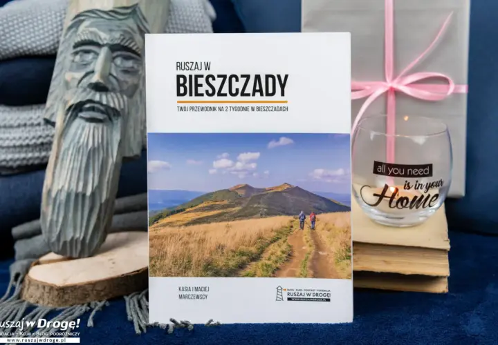Przewodnik "Ruszaj w Bieszczady" z widokiem na góry i ścieżkę, obok rzeźba i świeca.