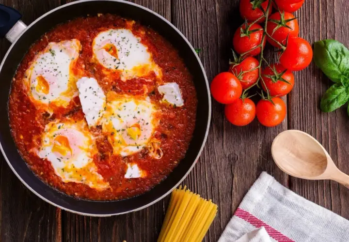 Uova al pomodoro in padella, con pomodorini freschi, basilico e un cucchiaio di legno, pronte per essere gustate.