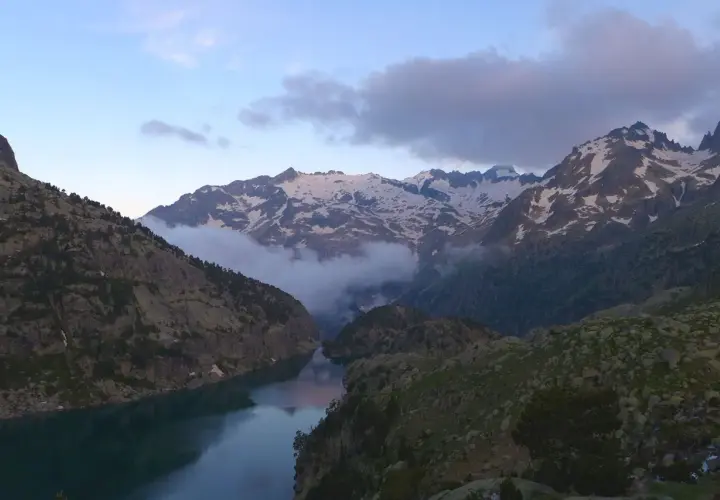Paisaje montañoso con un lago sereno y picos nevados. Nubes bajas envuelven las montañas, creando una atmósfera mística. Un lugar idílico para explorar, como el que se encuentra en Besiberri Sud.