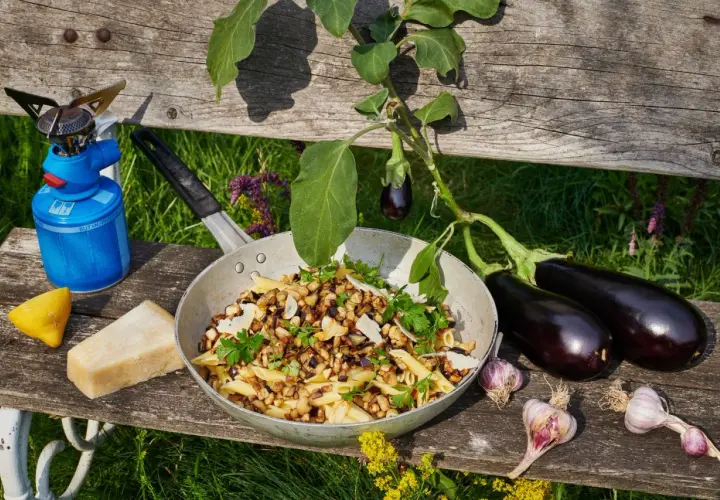 Pasta melanzane mit frischen Auberginen, Knoblauch und Parmesan. Ein Campingkocher und Zitrone deuten auf ein Outdoor-Mahl hin.