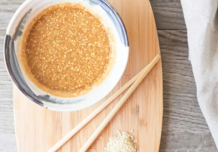 A bowl of creamy sesame sauce, perfect for your next sesame sauce recipe, sits on a wooden board with chopsticks and a sprinkle of sesame seeds.