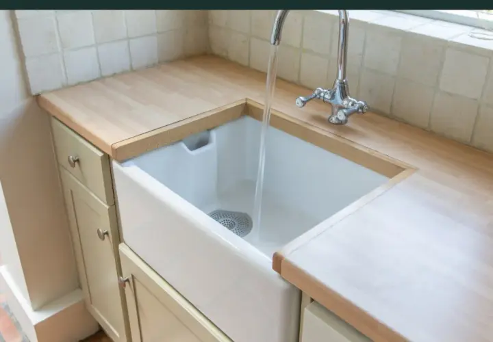 Biała, ceramiczna umywalka do kuchni z chromowaną baterią, w której płynie woda. Blat drewniany, szafki jasne.