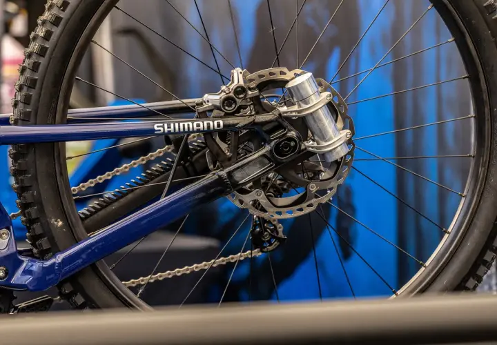 Close-up of a blue bicycle's rear wheel and Shimano drivetrain. A silver grip damper is visible on the axle, ready to absorb shocks.