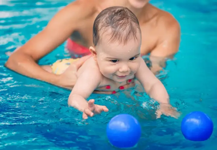 Glad bebis med ett leende i vattnet, en person håller i den. Två blå bollar flyter framför. Perfekt för babysim ålder.