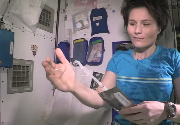 Astronautka pokazuje kroplę wody w dłoni, która unosi się w przestrzeni kosmicznej. Trzyma też opakowanie z napisem "Drinking Water".